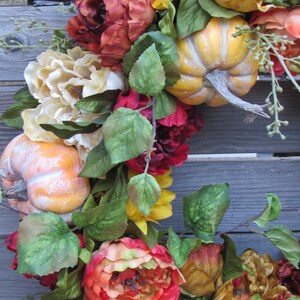 May include: A fall-themed wreath featuring red, orange and yellow flowers, green leaves and small pumpkins.