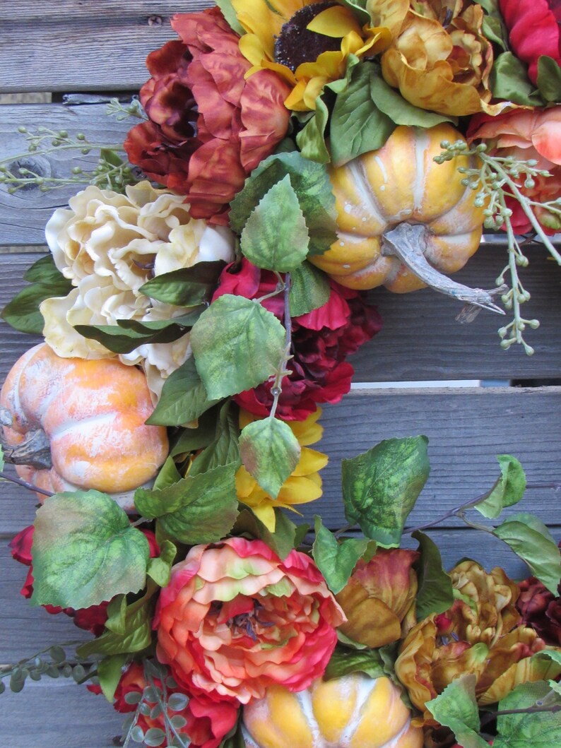 May include: A fall-themed wreath featuring red, orange and yellow flowers, green leaves and small pumpkins.
