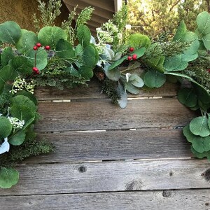 May include: A decorative garland featuring large, round green eucalyptus leaves, small red berries, and sprigs of evergreen. The garland is arranged on a weathered wooden surface, creating a natural and rustic aesthetic.