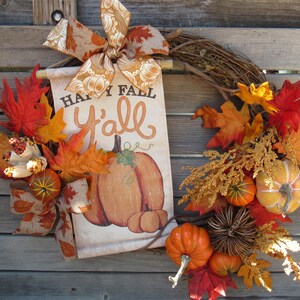 May include: A fall-themed wreath with a burlap bow, faux leaves, and pumpkins. A sign with the text "Happy Fall Y'all" is attached to the wreath.