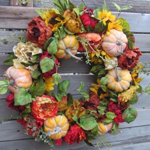 May include: A autumn-themed wreath featuring artificial sunflowers, red and orange peonies, and small orange pumpkins. The wreath is made on a grapevine base.