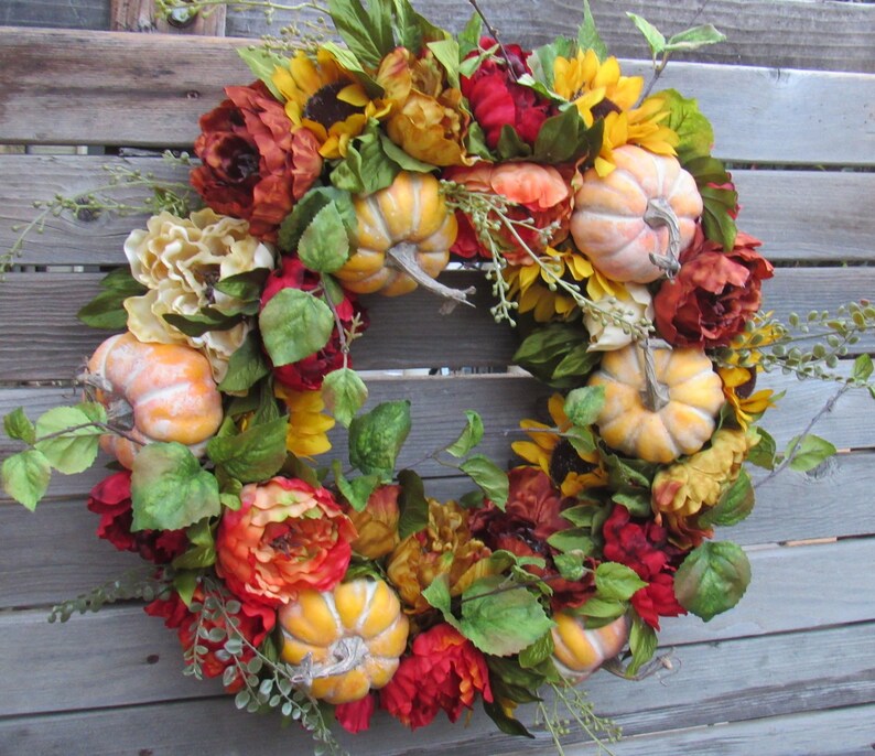 May include: A autumn-themed wreath featuring artificial sunflowers, red and orange peonies, and small orange pumpkins. The wreath is made on a grapevine base.