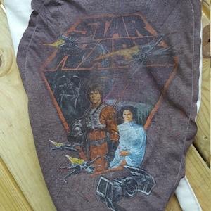 Op de afbeelding: Een bruine canvas draagtas met een Star Wars-afbeelding met Luke Skywalker, Prinses Leia en Darth Vader. De afbeelding is gedrukt op een bruin T-shirt dat op de tas is genaaid.