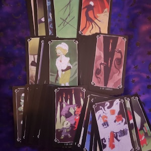 Pode incluir: Um baralho de cartas de tarot com ilustrações em estilo gótico. As cartas apresentam imagens de personagens e cenas, com uma borda preta e cantos decorativos. As cartas estão dispostas sobre uma superfície escura estampada.
