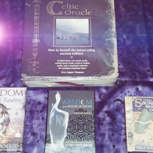 May include: Three different oracle card decks, including "Wisdom of the Hidden Realms", "Wisdom of the House of Night", and "The Sacred World Oracle". The decks are laid out on a purple fabric background. The book "Celtic Oracle" is visible in the background.