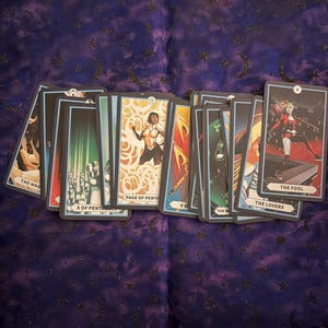 May include: A collection of tarot cards featuring colorful illustrations and titles like "The Fool" and "The Lovers." The cards are arranged on a dark purple cloth with a celestial pattern. Each card has a unique design and title.