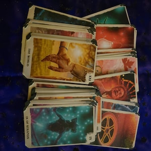 Könnte beinhalten: Ein Tarotkarten-Deck mit Vintage-Ästhetik. Die Karten zeigen Illustrationen verschiedener Figuren und Symbole, darunter eine ausgestreckte Hand, eine Glühbirne, ein Rad und eine Figur mit leuchtendem blauen Licht. Die Karten sind mit römischen Ziffern nummeriert, wie z. B. IX und XXI.