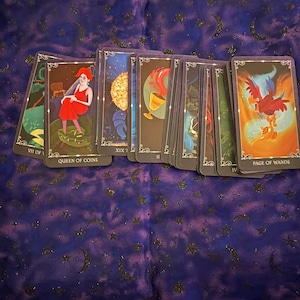 Puede incluir: Un conjunto de cartas del tarot con ilustraciones coloridas, incluyendo una mujer con un vestido rojo, un sol dorado y un pájaro. Las cartas están dispuestas sobre una tela morada oscura con un patrón celestial. El texto en las cartas incluye "QUEEN OF COINS" y "PAGE OF WANDS."