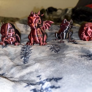 May include: Four small, red-toned figurines of fantasy creatures are arranged on a patterned surface. The figurines include a turtle-like creature, a dragon with wings, a frog, and a hedgehog. The figures are all approximately 2-3 inches tall.