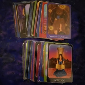 Może przedstawiać: Talia kart tarota z kolorowymi rewersami i różnymi ilustracjami. Jedna karta przedstawia kobietę czytającą książkę ze słowem "SOLITUDE" na dole. Inna karta wyświetla słowa "Obstacles & Challenges". Karty ułożone są na ciemnoniebieskiej powierzchni.