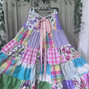 May include: A tiered maxi skirt featuring a patchwork design of floral, plaid, and gingham patterns in shades of pink, purple, green, blue, and yellow. The skirt has a high waist with a solid light pink fabric tie closure at the center front.