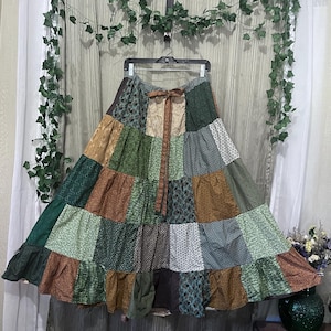 May include: A long, tiered patchwork skirt featuring a variety of fabric patterns in shades of forest green, olive, brown, and cream. The skirt has a gathered waist with a matching fabric tie and a wide, ruffled hemline at the bottom edge.