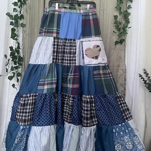 May include: A floor-length tiered skirt featuring a patchwork design of various blue, white, and green fabrics. The skirt includes sections of plaid, striped, and floral patterns, with a white square patch on the upper right side displaying a heart.