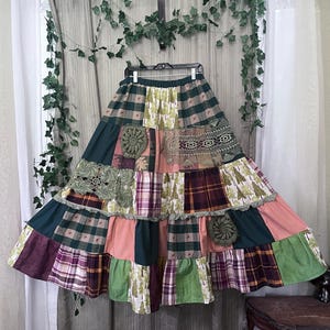 May include: A long, tiered patchwork skirt featuring a variety of fabric patterns including dark green, plaid, and tree-printed textiles. The skirt is embellished with several circular and rectangular pieces of olive green crochet lace trim.