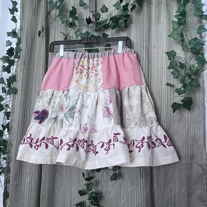 May include: A tiered skirt featuring a patchwork design with solid pink panels, floral print fabric, and a small heart-shaped patch. The bottom tier is made of cream-colored fabric with a dark pink embroidered floral vine pattern along the hemline.