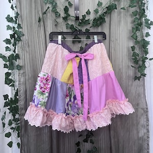 Puede incluir: Una falda de patchwork con varios paneles de tela en tonos rosa, morado, amarillo y estampados florales. La falda incluye un lazo rosa liso en la cintura y un dobladillo con volantes hecho de tela de encaje rosa claro con borde festoneado.