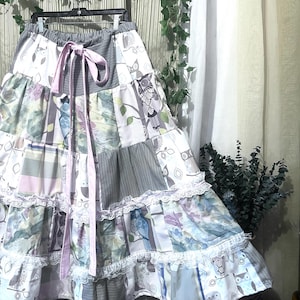 May include: A tiered maxi skirt featuring a patchwork design with owl illustrations, floral patterns, and geometric grey prints. The skirt includes a light pink ribbon drawstring at the elasticized waist and white lace trim accents between the fabric tiers.
