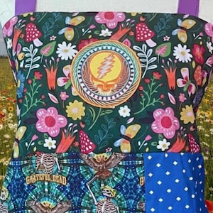 May include: An apron with a colorful floral pattern, mushrooms, and butterflies. The top section features a circular patch with a yellow lightning bolt and skull design. The bottom section has a blue tie-dye pattern with skeletons and the text "Grateful Dead".