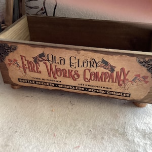 May include: A wooden box with the text "Old Glory Fire Works Company" in red and black. The box has a vintage design with illustrations of rockets and flags. The text includes "Bottle Rockets, Sparklers, Roman Candles".