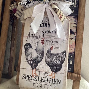 May include: A rustic wooden sign featuring a rooster and hen illustration, with the text "The Speckled Hen Farm." The sign is adorned with a decorative bow made of fabric strips and a daisy print. The sign has a distressed, vintage look.