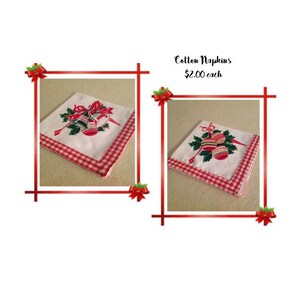 May include: Two white cotton napkins with red and green gingham trim. Each napkin features a Christmas bell design with red and gold accents. The text "Cotton Napkins &pound;2.00 each" is visible in the image.