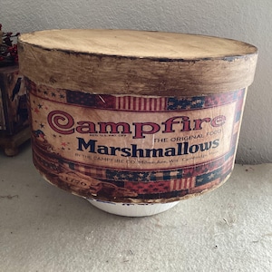 May include: A vintage Campfire Marshmallows container. The cylindrical container has a light brown lid and a body with a red, white, and blue patriotic design. The text "Campfire Marshmallows" is prominently displayed.