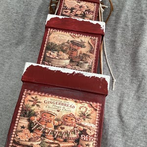 May include: A set of three stacked square wooden boxes with a distressed red finish and white painted edges. Each box features a vintage-style Christmas graphic with gingerbread characters and text including Wishing You Gingerbread Hugs.