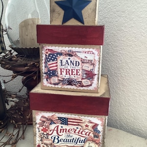 May include: Three stacked decorative boxes with red lids and patriotic designs. The top box has a blue star. The middle box reads "Land of the Free" and the bottom box says "America the Beautiful".