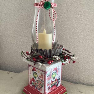 May include: A Christmas-themed candle holder with a clear glass hurricane lamp, a faux candle, and a decorative base. The base is red with green trim and features images of the Grinch with the text "Merry Grinchmas!"
