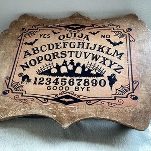 May include: A wooden Ouija board with a brown background and black lettering. The board features the letters A-Z, numbers 1-9, 0, and the words "YES", "NO", "GOOD BYE".