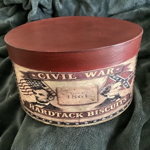 May include: A round, lidded container with a dark red top and a cylindrical body. The body features Civil War imagery, including the American flag, a Confederate flag, and portraits of historical figures. The text "CIVIL WAR HARDTACK BISCUITS SINCE 1861" is printed on the container.