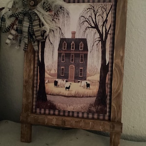 May include: Wooden framed farmhouse print with a pastoral scene. The print features a dark house, weeping trees, and sheep grazing in a field. The frame has a decorative plaid pattern and is accented with a fabric bow.