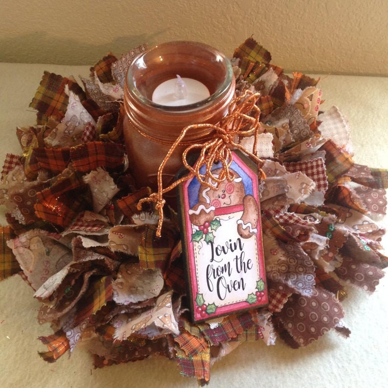May include: A brown and orange fabric wreath with a glass jar candle in the centre. The jar is decorated with copper paint and a brown ribbon. A gingerbread man tag with the text "Lovin' from the Oven" is attached to the jar.