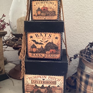 May include: A stack of three square black boxes with decorative labels. The top box reads PUMPKINS & CANDLELIGHT. The middle box reads BATS & BARN STARS. The bottom box reads PUMPKIN PATCH SISTERHOOD. Each box features rustic illustrations and tan twine.