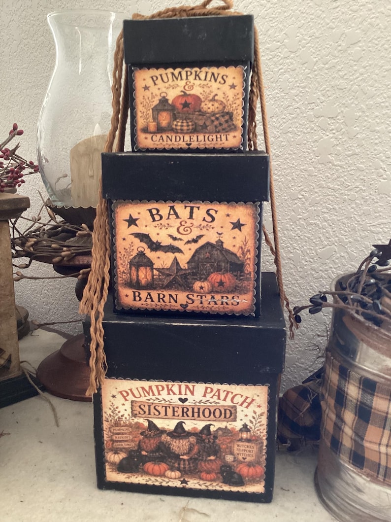 Peut inclure: Une pile de trois bo&icirc;tes carr&eacute;es noires avec des &eacute;tiquettes d&eacute;coratives. La bo&icirc;te sup&eacute;rieure indique PUMPKINS & CANDLELIGHT. La bo&icirc;te du milieu indique BATS & BARN STARS. La bo&icirc;te inf&eacute;rieure indique PUMPKIN PATCH SISTERHOOD. Ficelle beige.