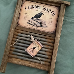 May include: A wooden washboard featuring a rectangular sign at the top with the text LAUNDRY SOAP CO. Est. 1887, a black crow illustration, and a stack of folded linens. A small matching tag is attached to the gray metal ridges with twine.