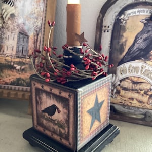 May include: A decorative candle holder with a black base and a square box design. The box features a crow illustration, a blue star, and a red berry garland. A faux candle with a star accent sits atop the box, creating a rustic, country aesthetic.