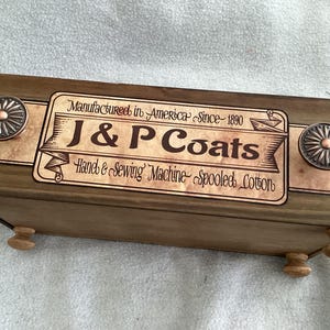 May include: A rectangular wooden storage box with a dark brown finish, featuring a vintage-style label that reads J & P Coats, Manufactured in America Since 1890, Hand & Sewing Machine Spooled Cotton. It has two round metal knobs and four small feet.
