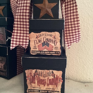 May include: A stack of three black boxes with patriotic labels and a rusty metal star on top. The boxes are adorned with labels from the "Old Glory Flag Company" and "Old Glory Fireworks Company". A red and white checkered ribbon is draped over the boxes.