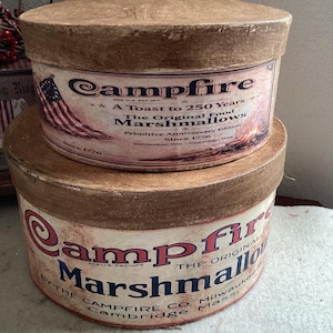 May include: Two vintage Campfire marshmallow tins. The tins are round with brown lids and sides. The labels feature the text "Campfire" in red and blue, with an American flag graphic. The text reads "A Toast to 250 Years".