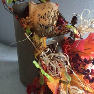 May include: A metal bucket filled with fall foliage, including red berries, orange and yellow leaves, and straw. A white candle with the word "gather" written on it is in the center of the arrangement.