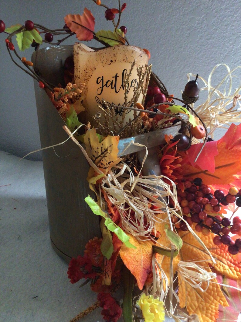 May include: A metal bucket filled with fall foliage, including red berries, orange and yellow leaves, and straw. A white candle with the word "gather" written on it is in the center of the arrangement.