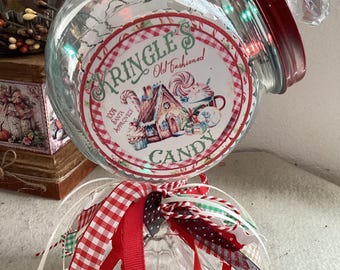 Könnte beinhalten: Ein klares Glasgefäß mit rotem Deckel und diamantförmigem Knauf, versehen mit einem runden Etikett mit der Aufschrift Kringle's Old Fashioned Candy 100% Santa Approved. Das Glas ist mit roten, weißen und grünen Bändern am Boden verziert.
