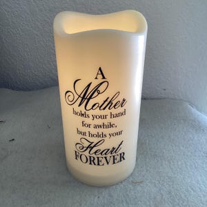 May include: A cream-colored candle with a wavy top and the text "A Mother holds your hand for awhile, but holds your Heart FOREVER" in black script. The candle is cylindrical and appears to be lit from within.