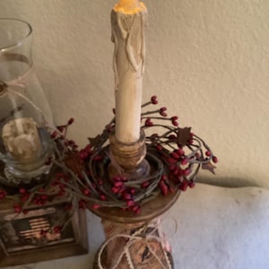 May include: A battery-operated candle with a flickering yellow flame sits atop a wooden candlestick. The candlestick is adorned with a red berry garland and a decorative base. A glass jar and a small wooden box are in the background.