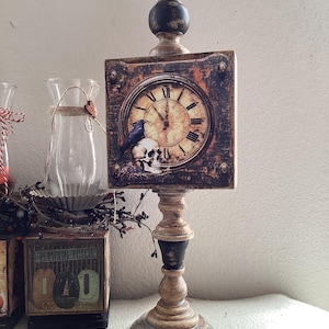 May include: A distressed wooden clock with a black raven and skull design. The clock is on a pedestal with a black and brown finish. The clock face has Roman numerals and a black hand.