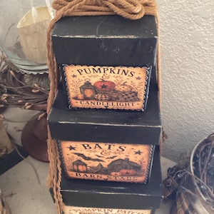 Peut inclure: Une pile de trois bo&icirc;tes carr&eacute;es noires attach&eacute;es avec une corde beige. La bo&icirc;te sup&eacute;rieure porte une &eacute;tiquette PUMPKINS & CANDLELIGHT. La bo&icirc;te du milieu porte une &eacute;tiquette BATS & BARN STARS. La bo&icirc;te inf&eacute;rieure porte PUMPKIN PATCH SISTERHOOD.
