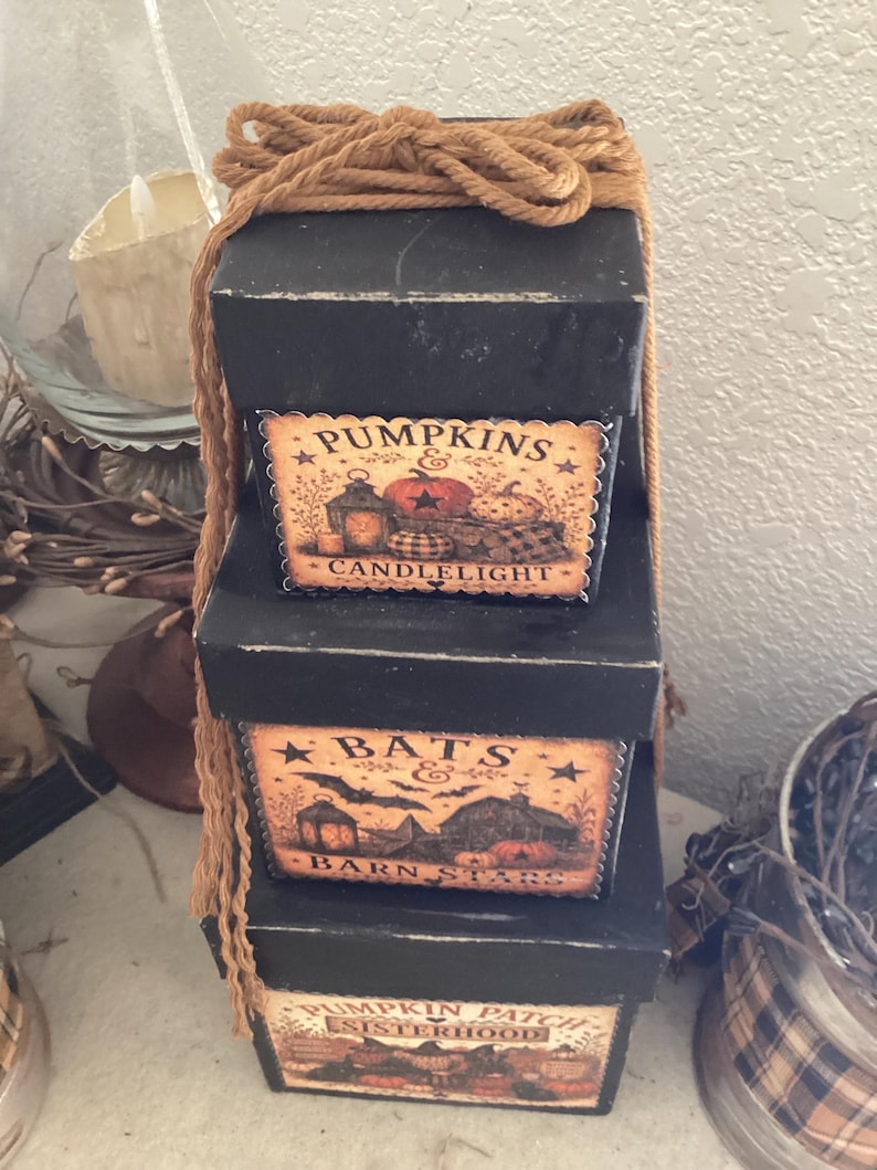 Peut inclure: Une pile de trois bo&icirc;tes carr&eacute;es noires attach&eacute;es avec une corde beige. La bo&icirc;te sup&eacute;rieure porte une &eacute;tiquette PUMPKINS & CANDLELIGHT. La bo&icirc;te du milieu porte une &eacute;tiquette BATS & BARN STARS. La bo&icirc;te inf&eacute;rieure porte PUMPKIN PATCH SISTERHOOD.