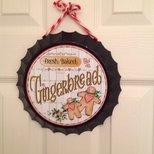 May include: A black metal sign with a white background and a red and white ribbon hanging from the top. The sign says "Fresh Baked Gingerbread" and features two gingerbread people.