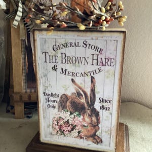 May include: A wooden sign for "The Brown Hare & Mercantile" with a rabbit illustration and text. The sign is topped with a candle and a decorative berry garland. The sign reads "Daylight Hours Only" and "Since 1892".